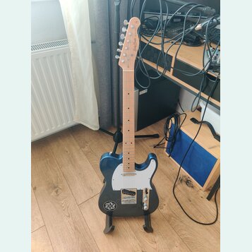 Modrá elektrická kytara s bílým pickguardem a světlou hnedou hmatníkovou deskou. Na hlavě je vidět nápis „Moody Blues“. Kytara stojí na černém stojan…