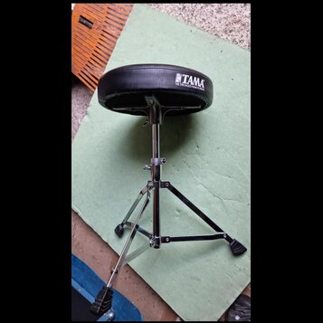 Černý bubeníkářský židle s kovovým chromovaným stativem a trojnožkou. Na sedáku je bílý nápis TAMA a pod ním text THE STRONGEST NAME IN DRUMS. Povrch…