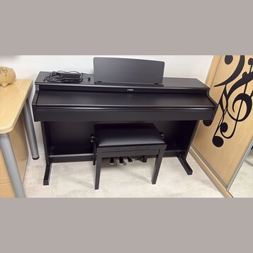 Černý digitální piano v černém nábytkovém stojanu s přívodním kabelem a doprovodnou černou stolečkou. Na přední straně je umístěno logo YAMAHA. Nástr…