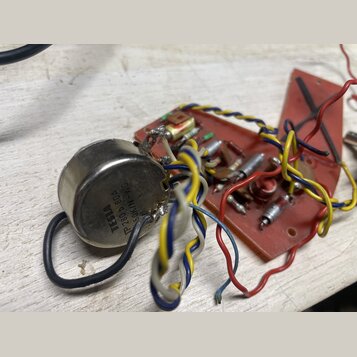 Fotografie zobrazuje elektronickou součástku v kovovém pouzdře připojenou k červené desce s dalšími komponenty a barevným zapojováním kabelů. Na kovo…