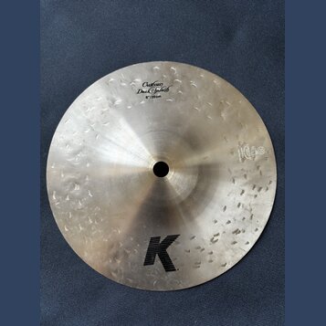 Kulatý činely z kovu s viditelným broušením povrchu. Na horní části je nápis „Custom Dark Splash 8"/20 cm“ a v dolní části velké černé logo „K“. V pr…