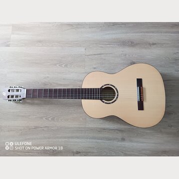 Kytara s přírodním dřevem na lícové desce a tmavým hnedým hmatníkem. Na hlavě nástroje je viditelné bílé logo v podobě stylizovaného písmena. Kytara…