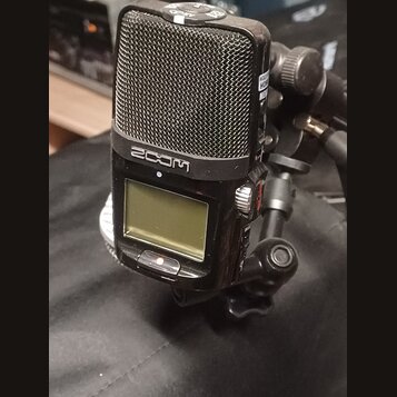 Černý digitální záznamový přístroj značky ZOOM s kovovou mřížkou mikrofonů v horní části. Zařízení má malý LCD displej a je připevněno na černý stati…