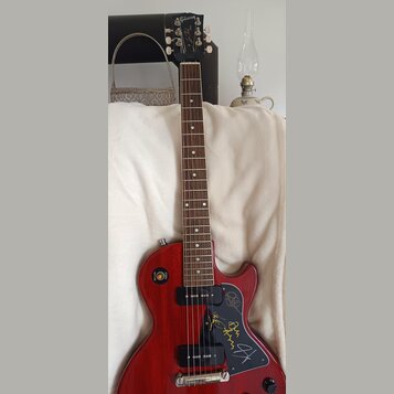 Červená elektrická kytara s tmavým hnedým hmatníkem. Na hlavě je bílé logo Gibson a nápis Les Paul. Černý plechtrguard obsahuje několik podpisů zlato…
