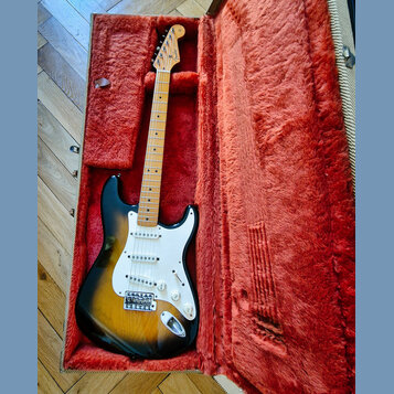 Elektrická kytara s tónem sunburst a bílým pickguardem v otevřeném kufru s červenou plišovou výstelkou. Na hlavě hředu je viditelné logo Fender. Kyta…