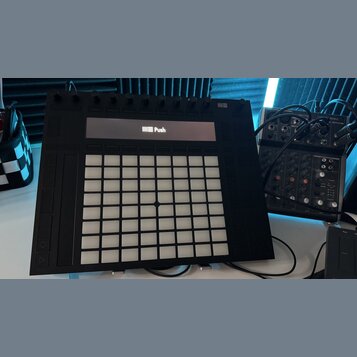 Černý kontrolér Ableton Push s mřížkou bílých podsvícených polí a řadou otočných ovladačů v horní části. Na displeji je vidět logo a nápis Push. Vedl…