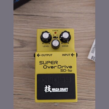 Žlutý kytarový efekt s nápisy SUPER Over Drive SD-1w a logem BOSS. Na horní části jsou tři bílé ovládací knoflíky s popisky LEVEL a DRIVE, přepínač S…