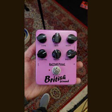 Růžový kytarový efekt s šesti černými ovládacími knoflíky. Na pedálu jsou nápisy LOW, MID, HIGH, LEVEL, VOICE a DRIVE, značka RAZZOR PEDAL. Ve spodní…