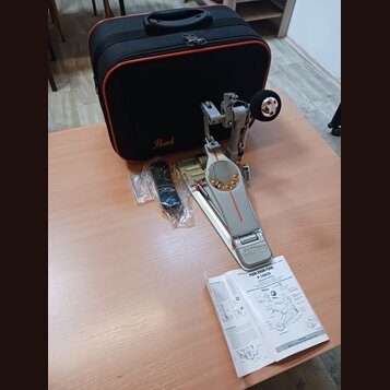 Na fotografii je stříbrný bubnový pedál s oranžovými detaily a nápisem IDemon. Vedle něj leží černý přepravní kufr s logem Pearl, náhradné části v pl…