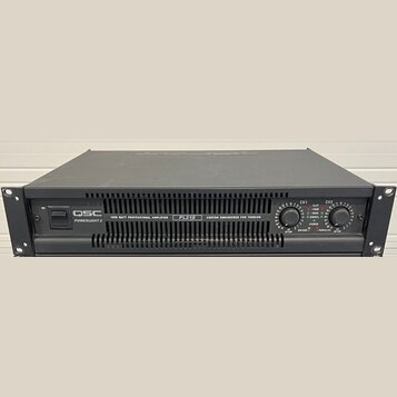 Černý rackový zesilovač s nápisy QSC POWERLIGHT 2, 1800 WATT PROFESSIONAL AMPLIFIER a PL218 CUSTOM ENGINEERED FOR TOURING. Na pravé straně jsou dva o…