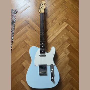 Světle modrá elektrická kytara typu Telecaster s bílým pickguardem a hnědým hmatníkem. Na hlavě nástroje je viditelné logo Fender. Kytara vykazuje mí…