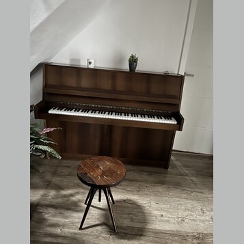 Hnědé dřevěné piano s klasickou klaviaturou. Před nástrojem stojí malá trojnožka dřevěná stolička s kulatým sedákem. Na horní desce piana jsou umístě…