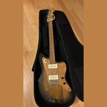 Elektrická kytara s přírodním dřevěným povrchem těla a hnedým hmatníkem, umístěná v černém obalu. Na hlavě je viditelné logo "Fender". Kytara má dva…