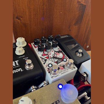 Detailní záběr na několik kytarových efektů v pedalboardu. Uprostřed je bílý pedál s červenobílou grafikou, který nese nápisy ratio, program a v doln…