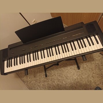Černý digitální klavír na černém stojanu. Na ovládacím panelu je bílý nápis Roland ep-7II DIGITAL PIANO. Nástroj má standardní klávesnici s bílými a…