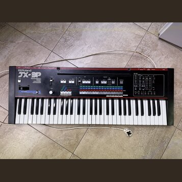 Černý syntetizátor s červeným horním panelem. Na levé straně je bílý nápis Roland a model JX-3P, pod kterým je text PROGRAMMABLE PRESET POLYPHONIC SY…