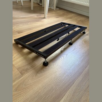 Černý kovový pedalboard s mřížkovým povrchem z gumových nebo plstěných pásů. Na boční hraně je bílý nápis „Harley Benton“ s logem. Nástroj stojí na č…