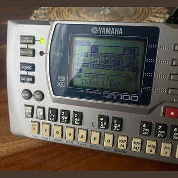 Stříbrný sekvenční modul s displejem a klávesovou ovládací. Na horní straně je logo YAMAHA a pod displejem nápis MUSIC SEQUENCER QY100. V levé části…