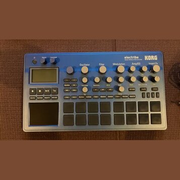 Modrý sekvenční syntetizátor s nápisy electribe MUSIC PRODUCTION STATION a KORG. Zařízení obsahuje displej, několik otočných knoflíků rozdělených do…