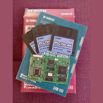 Fotografie zobrazuje červenobílý karton s nápisem YAMAHA PLG150-AN Plug-in Board, modrou obálku s logem YAMAHA a čtyři 3,5" diskety. Na disketách je…