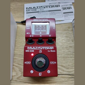 Červený efektový pedál s nápisy ZOOM a MULTISTOMP MS-60B for Bass. Zařízení má digitální displej, na kterém je vidět text DYNAMIC REVERB a tři ovláda…