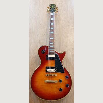 Elektrická kytara s tłem v barvě sunburst a zlatým hardwarem. Na hlavě je nápis johnson. Kytara má dva humbuckery, černý plechtrum a čtyři černé ovlá…
