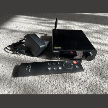 Černý audio přístroj s knoflíkem a anténou, na kterém jsou nápisy TOPPING a DX3Pro. Vedle přístroje je černý napájecí adaptér s kabelem a černý dálko…