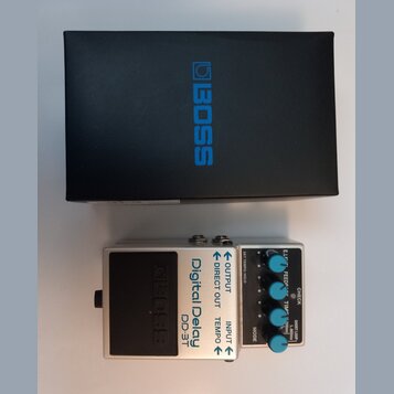 Na fotografii je stříbrný kytarový efekt s nápisy Digital Delay a DD-3T, umístěný před černým krabicem s modrým logem BOSS. Pedal má několik ovládací…