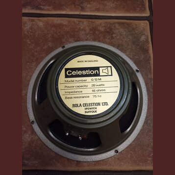 Kruhlový reproduktor s šedým rámem a žlutou štítkovou nálepkou. Na nálepce je uveden nápis Celestion, Model number G 12 M, Power capacity 25 watts, I…