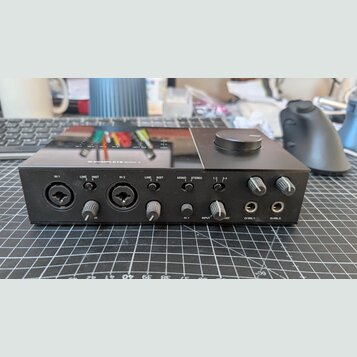 Černý zvukový interface s nápisem KOMPLETE AUDIO 6 na horní straně. Přední panel obsahuje dva kombinované vstupy (IN 1, IN 2), přepínače LINE/INST a…