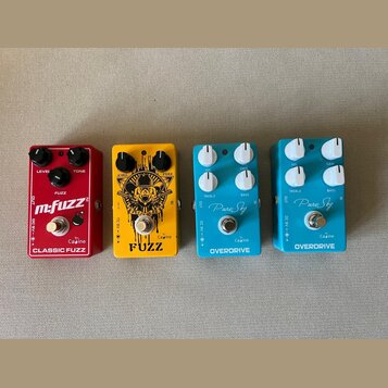 Čtyři kytarové efekty v řadě. Zleva: červený pedal s nápisy m:fuzz, Caïne a CLASSIC FUZZ; žlutý pedal s nápisem FUZZ a logem Caïne; modrý pedal s náp…