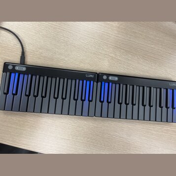 Černá elektronická klaviatura tvořená dvěma spojitými moduly. Na horní hraně obou částí je bílý nápis LUMI a ovládací tlačítka. Některé klávesy jsou…