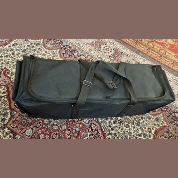 Černý textilní pouzdro (gig bag) obdélníkového tvaru s dvěma nosními popruhy. Povrch materiálu vykazuje drobné nečistoty a opotřebení. Na pouzdře nej…