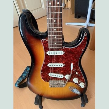 Elektrická kytara s tělem v barvě sunburst a hnědým hmatníkem. Nástroj má červený vzorovaný pickguard a tři bílé snímače. Na těle nejsou viditelné žá…