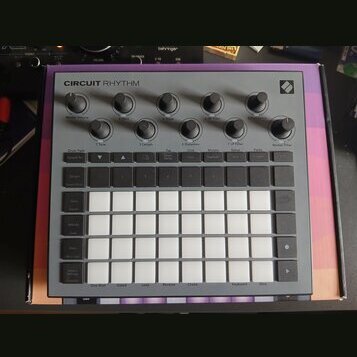 Šedý sekvenční sampler s názvem CIRCUIT RHYTHM a logem Novation v pravém horním rohu. Zařízení obsahuje několik otočných knoflíků, řadu malých tlačít…