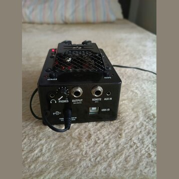 Černý elektronický přístroj s kovovou mřížkou na horní straně. Na zadní straně jsou viditelné nápisy BYPASS, DARK a konektory s popisky PHONES, OUTPU…
