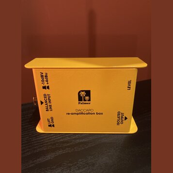 Žlutý kovový box s nápisy Palmer a DACCAPO re-amplification box. Na stranách jsou čitelné popisky BALANCED LINE INPUT s přepínačem -10dBV / +4dBu, LI…
