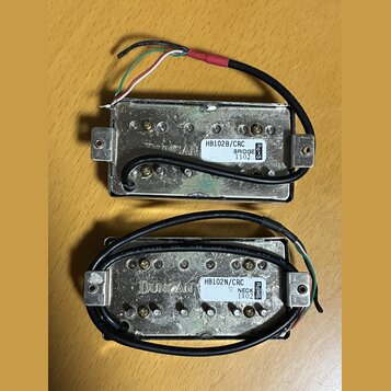 Dva kytarové humbuckery s kovovým krytem a připojenými kabely. Horní snímač má štítek s nápisy HB102B/CRC, BRIDGE a 1102. Dolní snímač má štítek s ná…