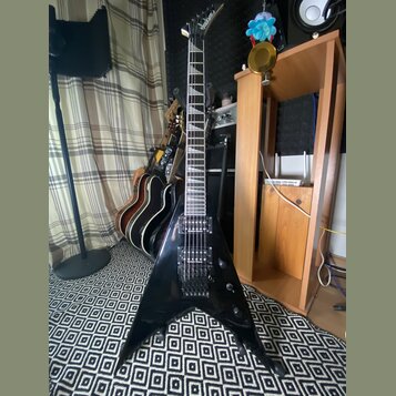 Černá elektrická kytara s tělem ve tvaru V. Na hlavě je viditelné logo Jackson. Kytara má dva humbuckery, fixační most a hриф s vkládanými značkami.…