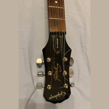 Detail hlavy černé kytary s hnedým hmatníkem. Na hlavě jsou bílé nápisy „GIBSON“, „Special MODEL“ a logo „Epiphone“. Jsou vidět šest chromovaných lad…