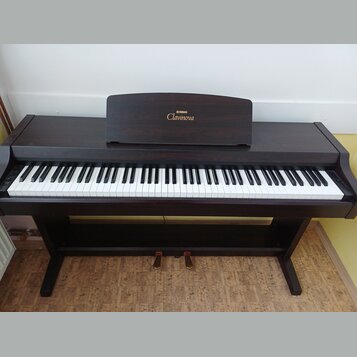 Digitální piano v tmavě hnědé barvě s dřevěnou strukturou. Na notovém pultu je zlatý nápis YAMAHA a Clavinova. Nástroj má standardní klávesnici s bíl…