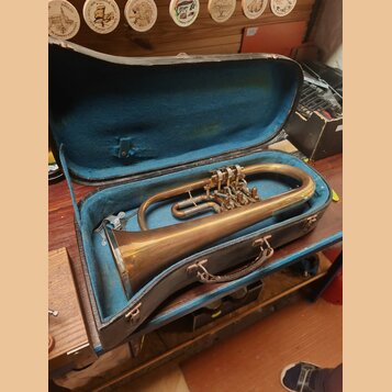 V otevřeném kufru s modrým vnitřkem leží mosazný žiletový nástroj (flugelhorn). Povrch kovu je matný s viditelnými oxidacemi a známky opotřebení. Na…