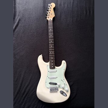 Bílá elektrická kytara typu Stratocaster s jasně zeleným pickguardem. Na hlavě hředu je viditelné logo Fender. Nástroj vykazuje známky používání, jso…