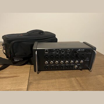 Černý digitální mixér s nápisem X AIR a logem behringer. Zařízení má řadu vstupních a výstupních konektorů s popisky jako REMOTE, ETHERNET, WIFI CLIE…