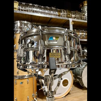Stříbrný kovový snare drum s chromovaným povrchem, umístěný na stojanu. Na korpusu je viditelné logo "Ludwig" v modrém obdélníku a žlutá nálepka s ná…
