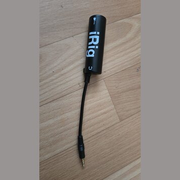 Černý cylindrický adaptér s nápisem iRig. Z přístroje vychází černý kabel zakončený 3,5mm jack konektorem. Povrch je matný a vykazuje mírné opotřeben…