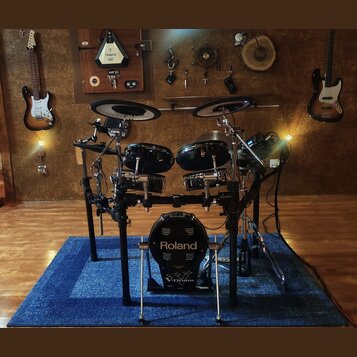 Elektronická bicí souprava značky Roland s černými bubny a činely. Na basovém bubnu je bílý nápis Roland a V-Drums. Souprava je umístěna na černém ko…