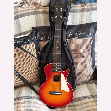 Červeno-žlutá akustická kytara s bílým plechtrem a černým popruhkem, opřená o černý pouzdro. Na hlavě nástroje je bílý nápis Epiphone a pod ním kurzi…