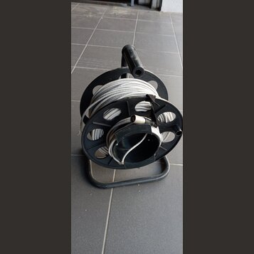 Černý plastový náviják s kovovým spodním rámem, na kterém je namotán šedý kabel. Na kabelu je viditelný konektor. Nástroj nemá žádné viditelné značky…
