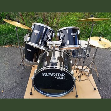 Černá bubnová sada s kovovým kováním a zlatými činely. Na basovém bubnu, tom-tamech i snare je viditelný nápis SWINGSTAR by TAMA. Na jednom z činelů…